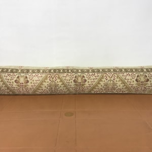 May include: A long, rectangular pillow with a beige background and a repeating geometric pattern in shades of brown and red. The pillow is placed on a brown surface, and the background is white.