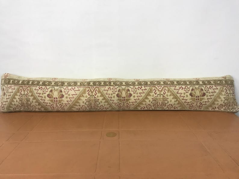 May include: A long, rectangular pillow with a beige background and a repeating geometric pattern in shades of brown and red. The pillow is placed on a brown surface, and the background is white.