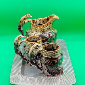 May include: A brown and white ceramic pitcher and three mugs with dog-shaped handles, all sitting on a silver tray with a textured surface.