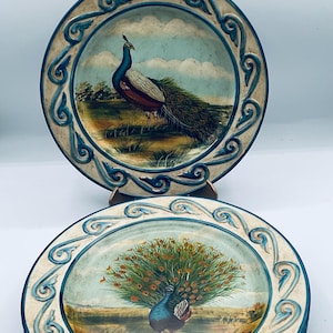 May include: Decorative plates featuring peacock designs. The plates have a cream-colored border with a blue scroll design. The peacock illustrations are set against a landscape with a blue sky.