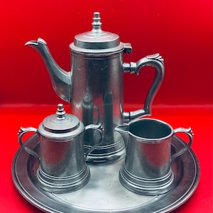 May include: A pewter tea set consisting of a tall teapot with a lid, a lidded sugar bowl, a creamer, and a round serving tray. Each piece features a smooth, metallic gray finish with a simple, functional design and curved handles.
