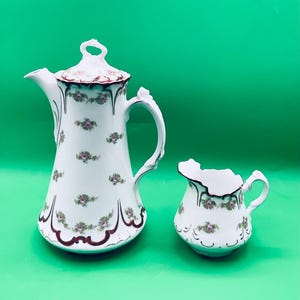 May include: A white porcelain teapot and creamer set with a floral pattern in shades of pink and green. The teapot has a lid with a decorative knob and a spout. The creamer has a handle and a rounded shape.