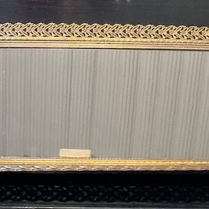May include: A gold-colored metal tray with a mirrored surface and a decorative, ornate border. The tray has a rectangular shape with slightly rounded corners.