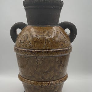May include: A decorative ceramic vase with a textured, aged appearance. The vase features a dark brown rim and handles, with a lighter brown body. The body has horizontal bands and a cracked, weathered effect, suggesting an antique or rustic style.