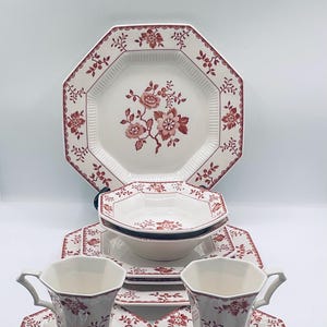 May include: A white ceramic dinnerware set with a red floral pattern. The set includes a large octagonal plate, bowls, smaller plates, teacups, and saucers. The plates and cups have a scalloped edge.