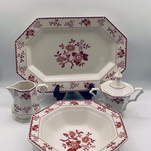 May include: A vintage white porcelain dish set with a floral pattern in shades of red. The set includes a serving platter, a bowl, a creamer, and a sugar bowl with a lid. The edges of the pieces have a scalloped design.