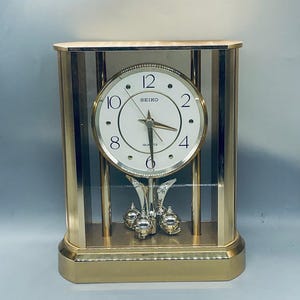 May include: A gold-toned Seiko quartz mantel clock with a pendulum and three swinging balls. The clock face is white with black Roman numeral hour markers and black hands.