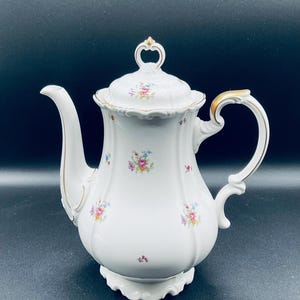May include: White porcelain coffee pot with a gold trim and floral pattern. The pot has a lid with a gold knob and a curved handle.