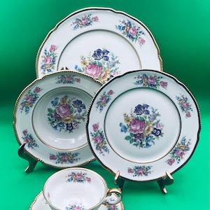 May include: A vintage floral china set featuring a teacup and saucer, a bowl, and two plates. The white porcelain pieces have a gold trim and are decorated with a colorful floral pattern of pink, purple, and yellow flowers.
