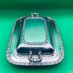 May include: A silver-colored, rectangular serving dish with a lid. The dish has ornate, scrolled detailing around the edges and handles. The lid has a recessed center and a handle. The background is a solid green.