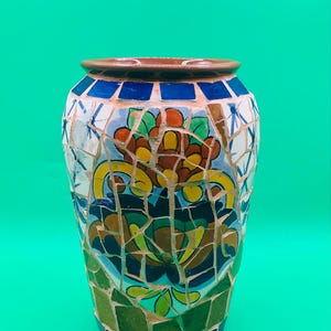 May include: A brown ceramic vase with a mosaic design featuring blue, green, yellow, red, and white tiles. The vase has a floral pattern with a large flower in the center.