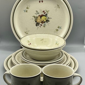 May include: A set of vintage dinnerware featuring a cream-colored background with a green and brown border. The design includes a cluster of fruit, including pears, apples, and berries, with green leaves. The set includes a large dinner plate, a smaller salad plate, a bowl, and two mugs.
