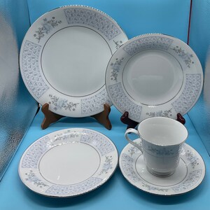 May include: A set of white porcelain dinnerware with a delicate blue and gray floral pattern. The set includes a dinner plate, a salad plate, a soup bowl, and a teacup and saucer.