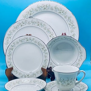 May include: A set of white porcelain dinnerware with a delicate floral pattern in shades of green and gray. The set includes a large dinner plate, a salad plate, a soup bowl, a teacup, and a saucer.