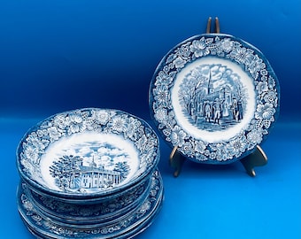 Puede incluir: Un conjunto de platos y cuencos de cerámica azul y blanca. Los platos y cuencos presentan un borde floral y una imagen central de un edificio o iglesia. Un plato se exhibe en un soporte dorado. Los artículos están dispuestos sobre un fondo azul.