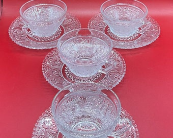 May include: Five clear glass teacups and saucers with ornate, embossed floral and geometric patterns. The cups and saucers are arranged on a red background. The teacups have handles and are filled with a clear liquid.