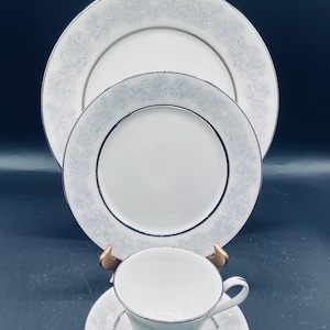 May include: A set of white porcelain dinnerware with a delicate silver trim and a floral pattern. The set includes a large dinner plate, a smaller salad plate, and a teacup and saucer.