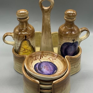 May include: Ceramic olive oil and vinegar set with a handle. The set features two bottles with painted pears and plums, and a small dish with a painted plum. The set is brown and beige.