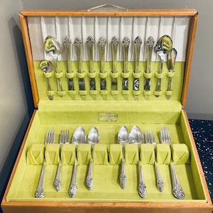 May include: A wooden box with a green velvet lining containing a set of silverware. The silverware is arranged in rows and includes forks, knives, spoons, and a butter knife. The box has a label that reads "1881 Rogers Co."