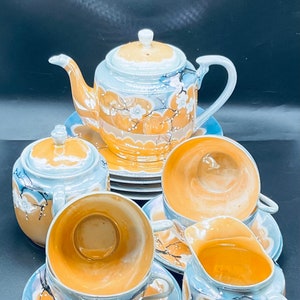 May include: A vintage Japanese tea set with a floral design. The set includes a teapot, creamer, sugar bowl, and two teacups and saucers. The teacups and saucers have a light blue and orange color scheme with a floral design.
