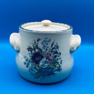 May include: White ceramic crock with a lid and two handles. The crock has a floral design with blue, purple, and green flowers.