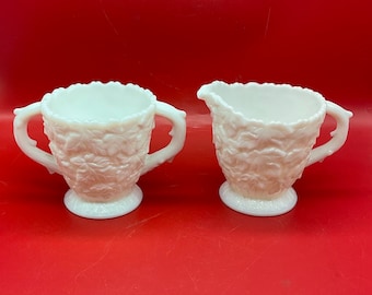 May include: A white milk glass creamer and sugar bowl set with a leaf design. The creamer has a handle and a spout. The sugar bowl has two handles.
