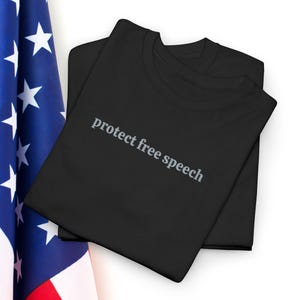 Puede incluir: Dos camisetas negras apiladas, con la frase "protect free speech" impresa en gris. Las camisetas están colocadas junto a una porción de la bandera estadounidense, con su campo azul y estrellas blancas.