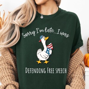 May include: Dark green t-shirt with a cartoon goose wearing a patriotic hat and vest. The text on the shirt reads "Sorry I'm late...I was Defending Free Speech". The goose has yellow feet and a yellow beak. The t-shirt is a casual style.