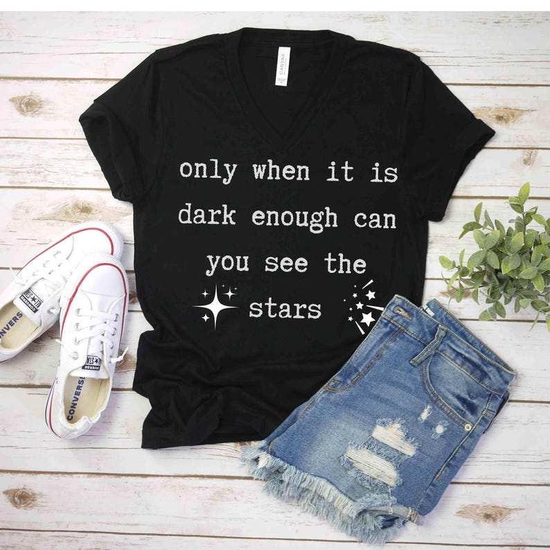 Op de afbeelding: Zwart V-hals T-shirt met de tekst "only when it is dark enough can you see the stars" in wit. Het shirt is gecombineerd met een spijkerbroek.