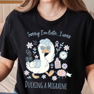 Puede incluir: Camiseta negra con un pato de dibujos animados con gafas de sol y una taza, con el texto "Sorry I'm late...I was ducking a migraine." El diseño incluye flores, pastillas y un reloj.