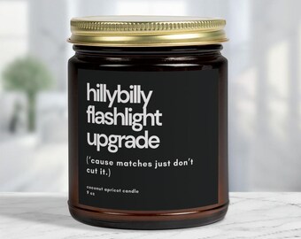 Puede incluir: Un tarro de vela de vidrio marrón con tapa dorada. La etiqueta dice "hillybilly flashlight upgrade ('cause matches just don't cut it.) coconut apricot candle 9 oz".