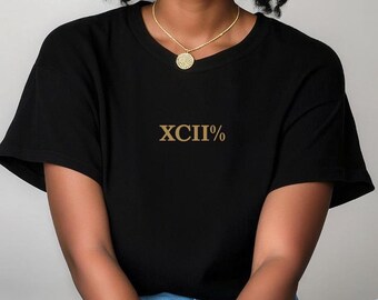Op de afbeelding: Een zwart T-shirt met een gouden Romeins cijfer 'XCII' en een procentteken. Het shirt wordt gedragen door een persoon met krullend haar en gouden sieraden.