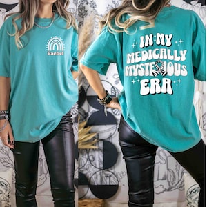 May include: A turquoise blue oversized t-shirt with a rainbow graphic and the name "Rachel" on the front. The back of the shirt has the text "In My Medically Mysterious Era" with a zebra print graphic.