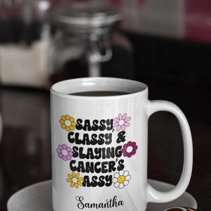 May include: White ceramic coffee mug with a black design that says "Sassy, Classy & Slaying Cancer's Ass" with a daisy pattern around the text. The mug has a black handle and the name Samantha is written at the bottom.