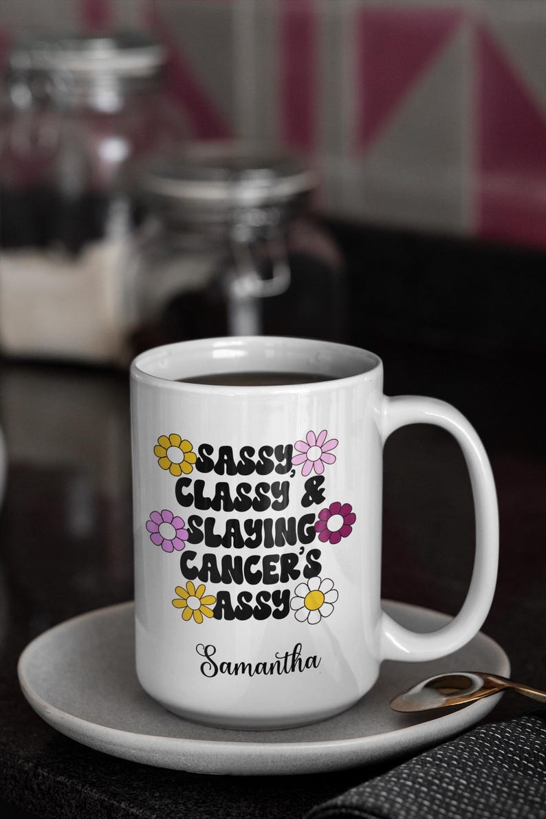 May include: White ceramic coffee mug with a black design that says "Sassy, Classy & Slaying Cancer's Ass" with a daisy pattern around the text. The mug has a black handle and the name Samantha is written at the bottom.