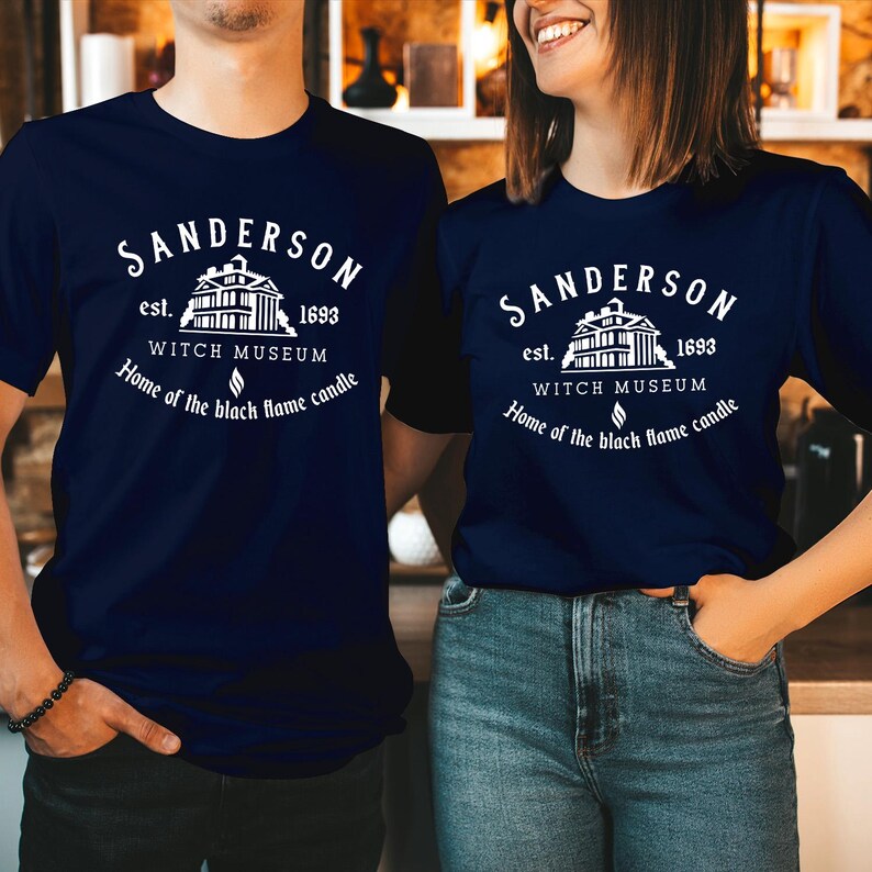 May include: A navy blue t-shirt featuring a white graphic print that reads Sanderson Witch Museum est. 1693, Home of the black flame candle, accompanied by an illustration of a building and a small flame icon centered on the chest.