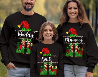 May include: Three black long-sleeve sweatshirts featuring a Christmas elf graphic with a red hat, striped legs, green shoes, and gift boxes. The shirts display the text Daddy, Mummy, and Emily, accented by a string of colorful holiday lights.