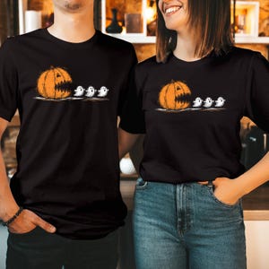 May include: A black short-sleeved t-shirt featuring a graphic print of an orange jack-o'-lantern with a wide, jagged mouth chasing three small white ghosts in a line. The design is centred on the chest area of the garment.