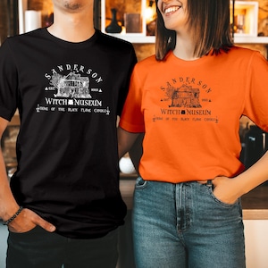 May include: A black t-shirt and an orange t-shirt, each featuring a white or black graphic print of a house with the text Sanderson Witch Museum, Est. 1693, Home of the Black Flame Candle, and a small cauldron icon centered below the house.