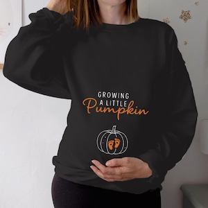 Puede incluir: Una sudadera negra de manga larga con el texto en blanco y naranja "GROWING A LITTLE Pumpkin" sobre un gráfico de calabaza en líneas blancas. La calabaza contiene dos pequeñas huellas de bebé naranjas con talones en forma de corazón.
