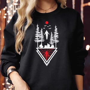 May include: Black sweatshirt with a white and red graphic design. The graphic features a stylized forest scene with figures, trees, and a red sun. The design is within a white triangle.