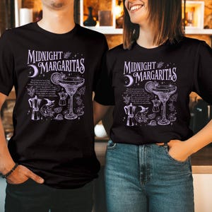 May include: A black t-shirt featuring a light purple graphic print with the text "MIDNIGHT MARGARITAS". The design includes a margarita glass, a frog wearing a witch hat, a coffee maker, a snail, mushrooms, and a crescent moon with stars.