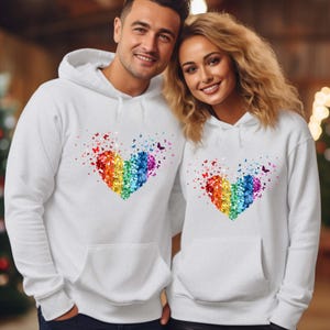 Puede incluir: Sudaderas con capucha blancas con un diseño de corazón arcoíris hecho de mariposas de colores. Las sudaderas tienen un bolsillo delantero y una capucha. La imagen muestra a dos personas usando las sudaderas.