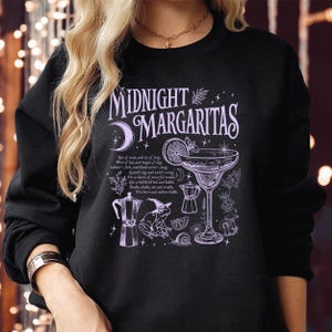 May include: A black long-sleeve sweatshirt featuring a light purple graphic print that reads Midnight Margaritas. The design includes a margarita glass, a frog wearing a witch hat, two moka pots, a crescent moon, and small text excerpts.