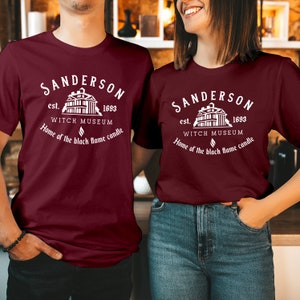May include: A maroon short-sleeved t-shirt featuring a white graphic print that reads: SANDERSON est. 1693 WITCH MUSEUM Home of the black flame candle, accompanied by an illustration of a building and a small flame icon centered above the text.