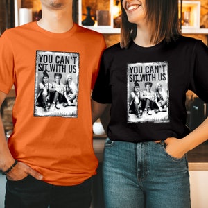 May include: An orange t-shirt and a black t-shirt, each featuring a rectangular graphic print of three figures in witch hats sitting together with the text "YOU CAN'T SIT WITH US" printed in bold black capital letters above the figures.