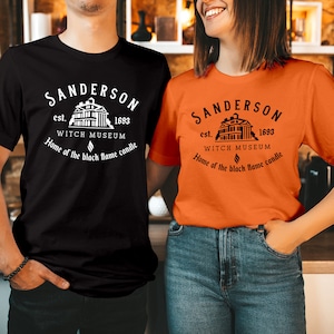 May include: A black t-shirt and an orange t-shirt, each featuring a white or black graphic print that reads: Sanderson est. 1693 Witch Museum, Home of the black flame candle, with an illustration of a building and a small flame icon centered.
