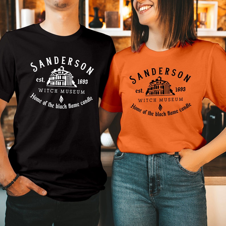 May include: A black t-shirt and an orange t-shirt, each featuring a white or black graphic print that reads: Sanderson est. 1693 Witch Museum, Home of the black flame candle, with an illustration of a building and a small flame icon centered.