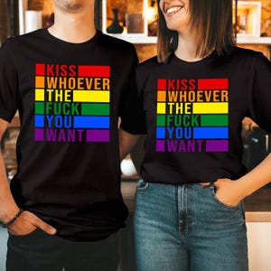May include: Black t-shirts with a rainbow-colored text design that reads "KISS WHOEVER THE FUCK YOU WANT." The text is arranged in horizontal bands of red, orange, yellow, green, blue, and purple. The shirts are worn by two people.