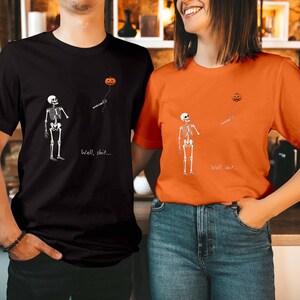 May include: A black t-shirt and an orange t-shirt, each featuring a white skeleton graphic looking up at a floating jack-o'-lantern balloon on a string. Below the skeleton, the text "Well, shit..." is printed in a simple white font.